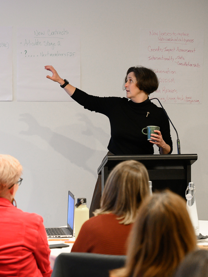 An image of Helen wearing a black, high necked top and holding a green mug. She is standing behind a lectern and gesturing at a white board.