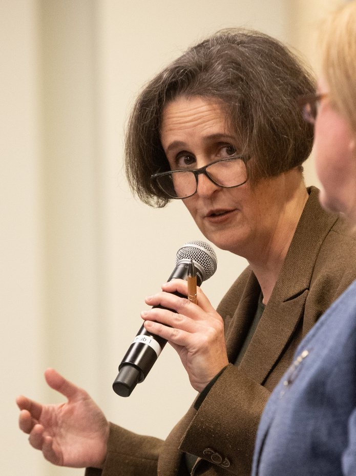 An image of Helen wearing a brown jacket. She is taking into a hand held microphone.