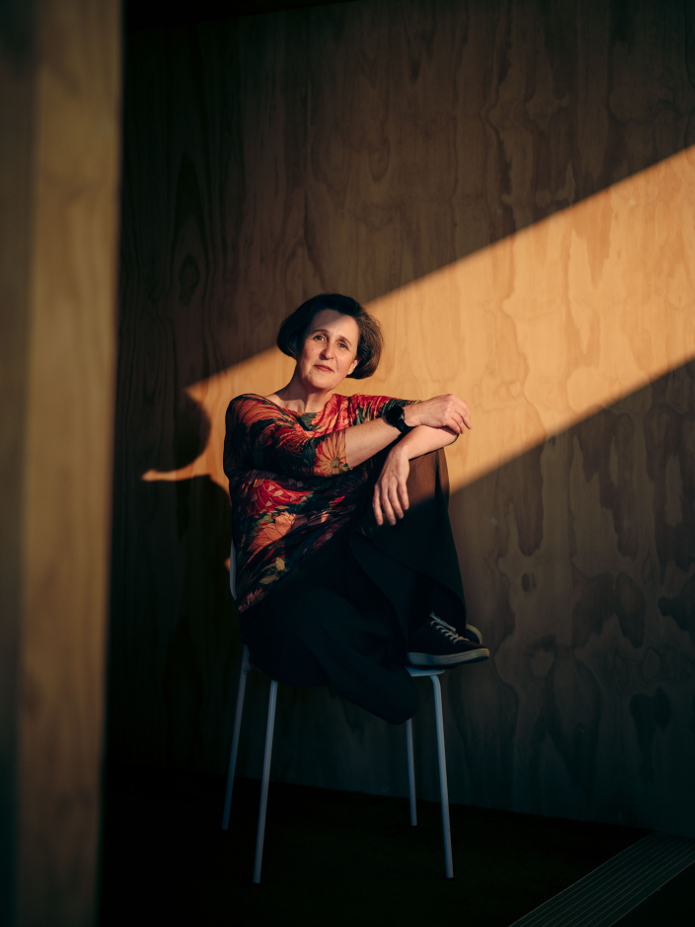 An image of Helen, who is a white middle aged woman seated on a chair. her face is lit by a dramatic line of light, but her body is in shadow.