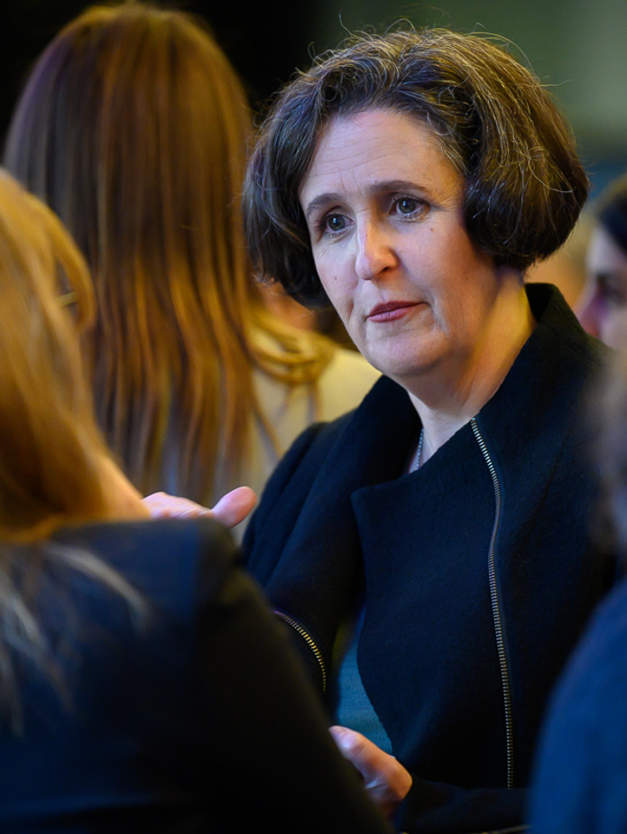 An image of Helen wearing a black zip up jacket. She is at a function in a crowd of people listening to another woman talk.